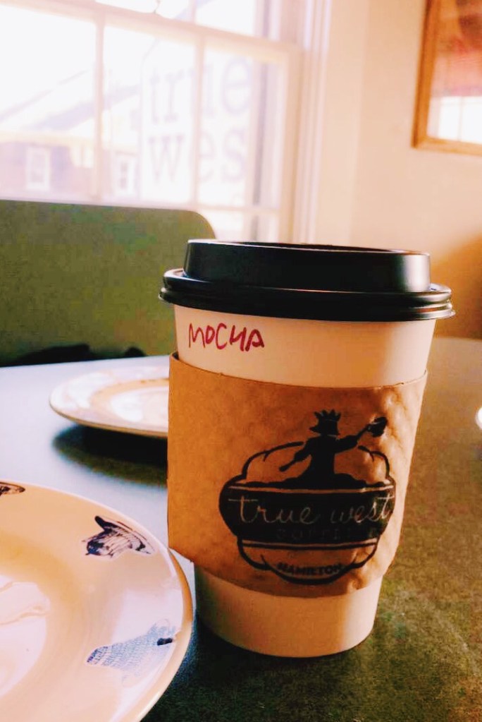 A coffee cup sitting on a table. It has the True West Coffee logo and also has "mocha" written on it with permanent marker