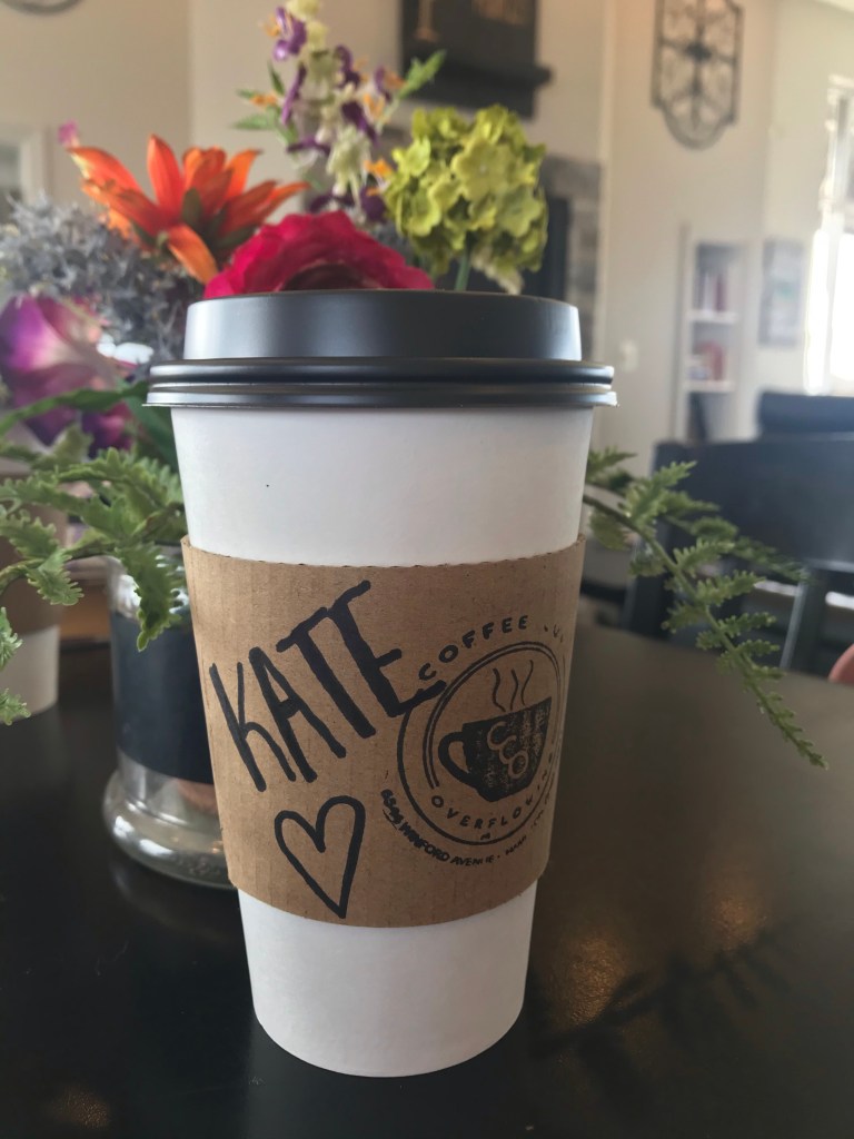 A coffee cup that says "Coffee Cup Overflowing" and has the name, "Kate" written with a marker. It is sitting next to a vase of flowers