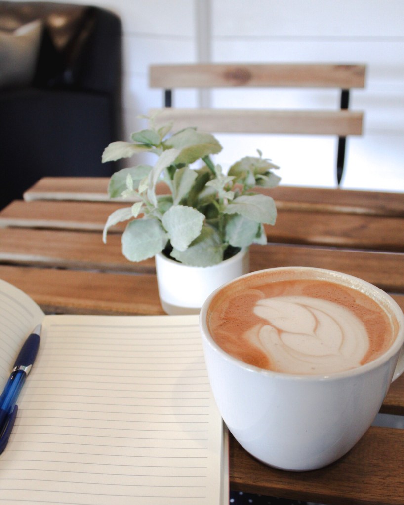 Photo of a latte in a mug on top of a wooden table. It is next to a plant and a journal.